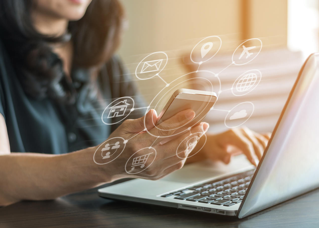 A woman is seated with her laptop open and is multitasking by using her phone to execute digital marketing strategies online
